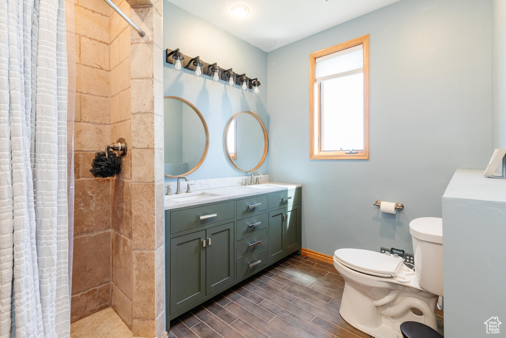 Full bath featuring toilet, double vanity, tiled shower, and wood finish floors