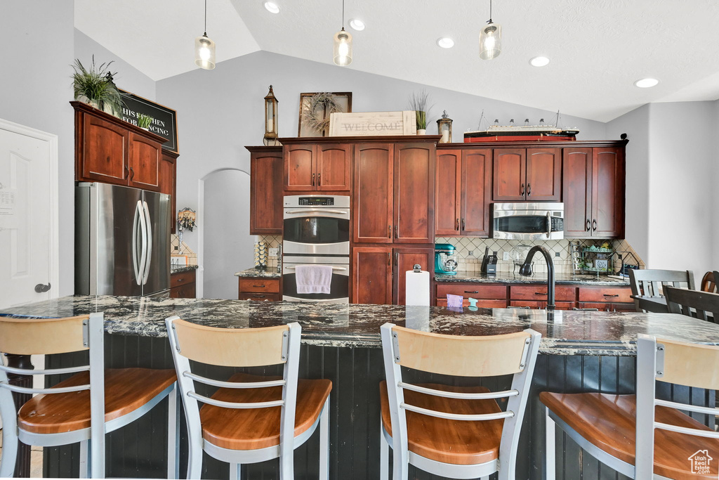Kitchen featuring appliances with stainless steel finishes, vaulted ceiling, reddish brown cabinets, decorative backsplash, and pendant lighting