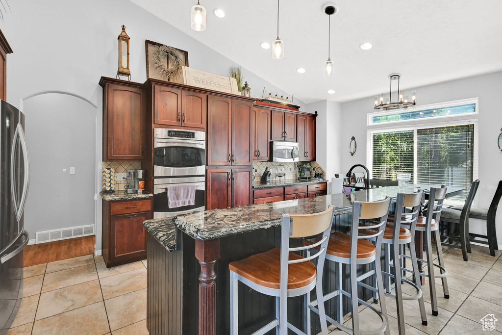 Kitchen with stainless steel appliances, tasteful backsplash, light tile patterned flooring, a center island with sink, and decorative light fixtures