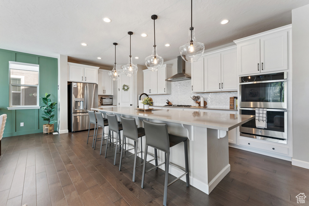 Kitchen featuring stainless steel appliances, wall chimney exhaust hood, tasteful backsplash, dark wood finished floors, and white cabinets
