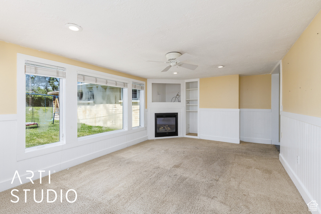 Unfurnished living room featuring a ceiling fan, a wainscoted wall, carpet, healthy amount of natural light, and a glass covered fireplace
