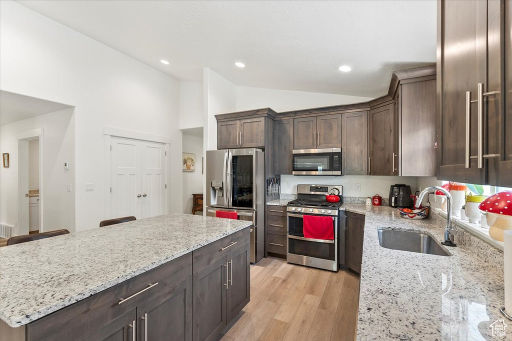 Kitchen featuring stainless steel appliances, a sink, vaulted ceiling, dark brown cabinets, and light stone countertops