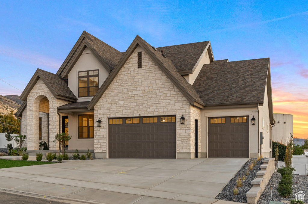 View of front facade with an attached garage, stone siding, driveway, stucco siding, and roof with shingles