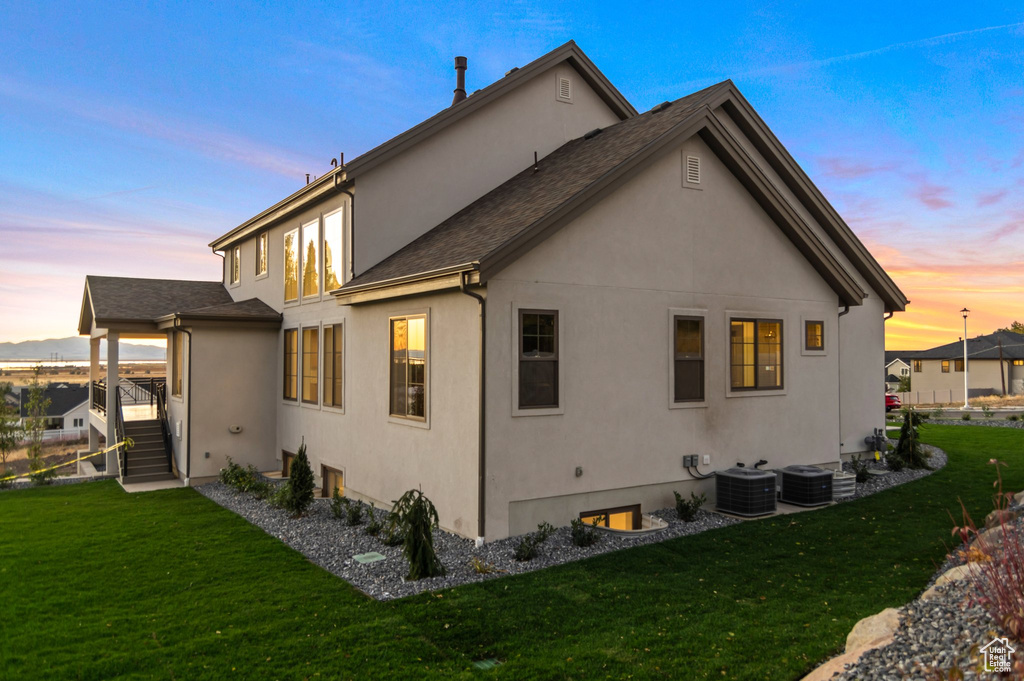 Back of house at dusk with stucco siding, a lawn, central air condition unit, stairway, and a shingled roof