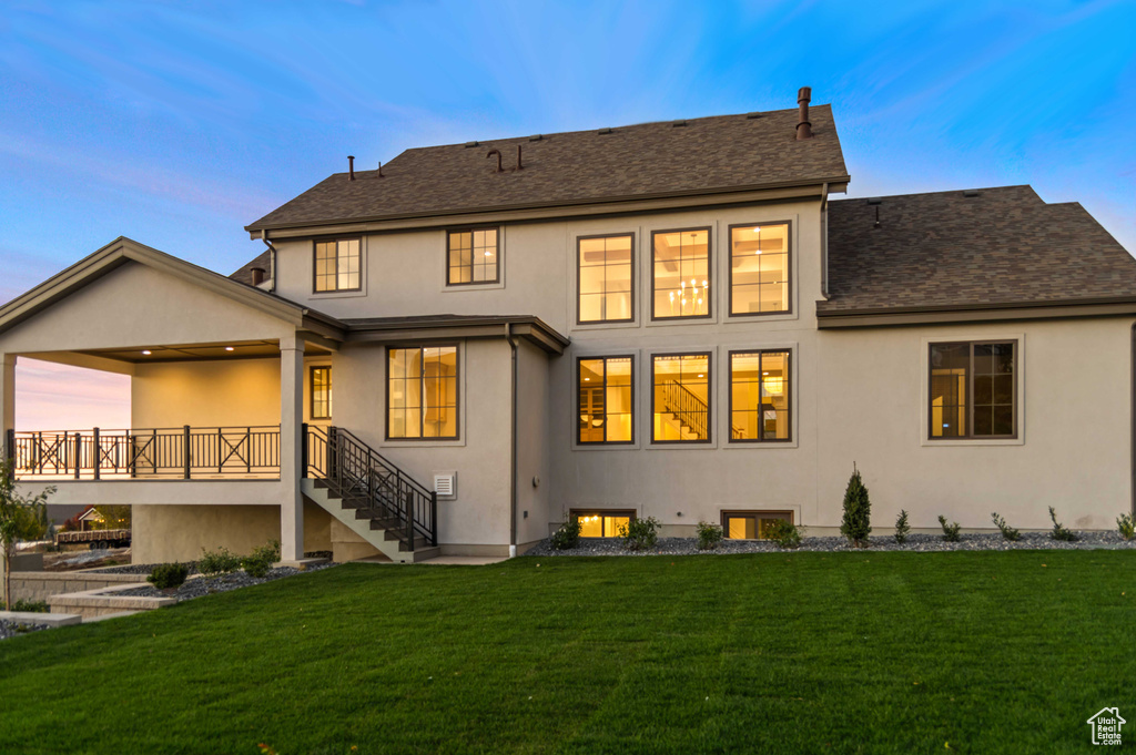 Back of property at dusk with stucco siding, a yard, stairs, and roof with shingles