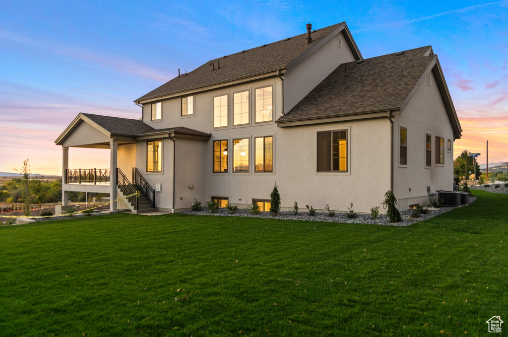 Back of house at dusk with a yard, stucco siding, central AC unit, and a shingled roof