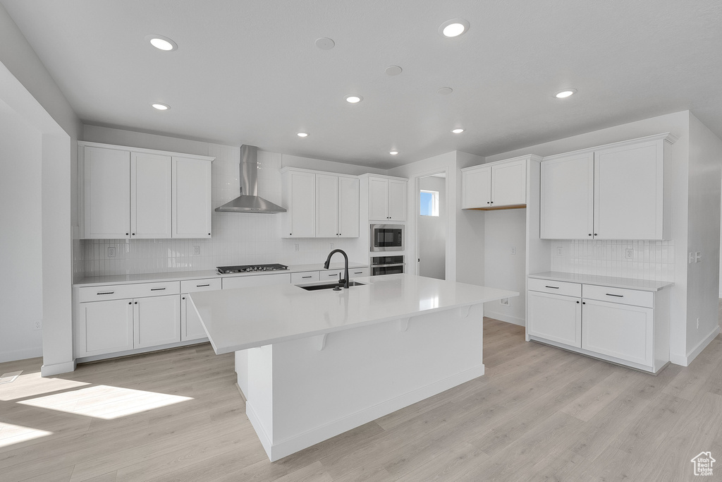 Kitchen featuring wall chimney exhaust hood, a sink, appliances with stainless steel finishes, an island with sink, and decorative backsplash