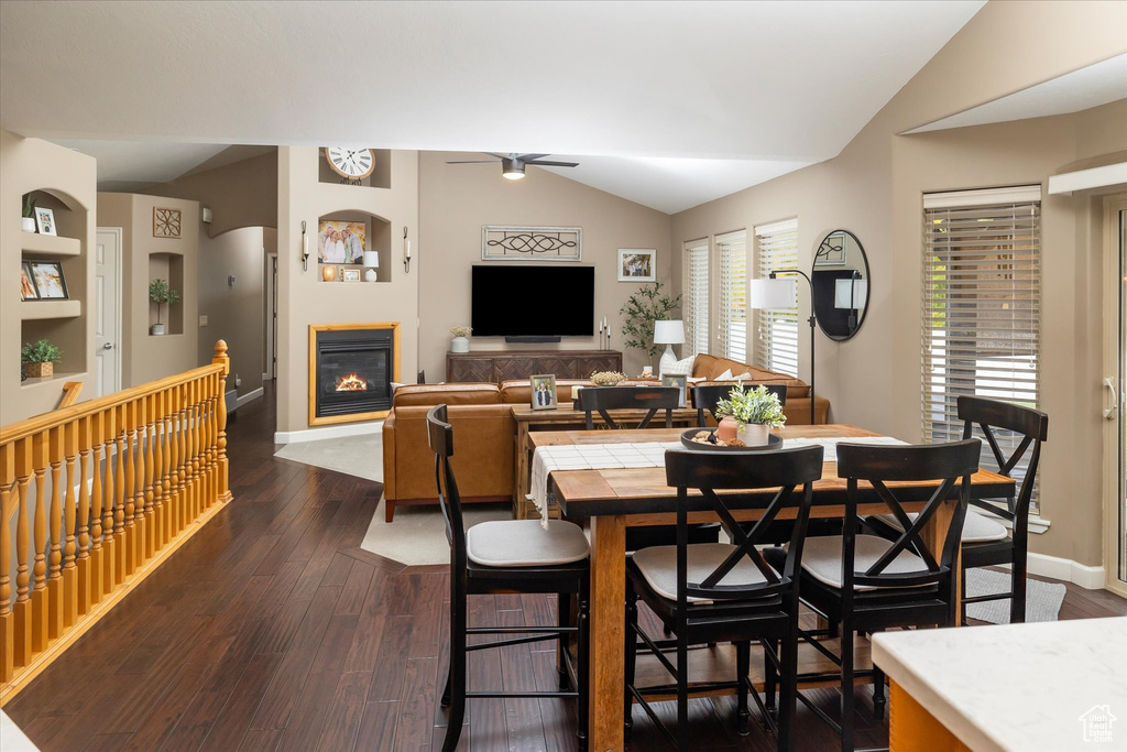 Dining area with vaulted ceiling, a ceiling fan, a glass covered fireplace, dark wood-style flooring, and baseboards