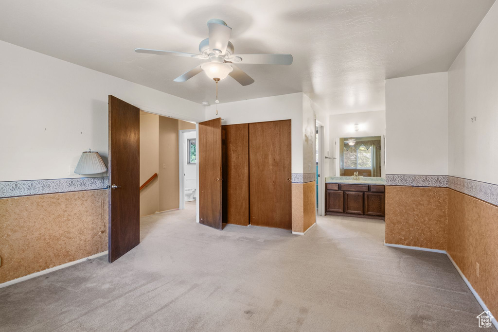 Unfurnished bedroom with wainscoting, light colored carpet, ensuite bath, and ceiling fan