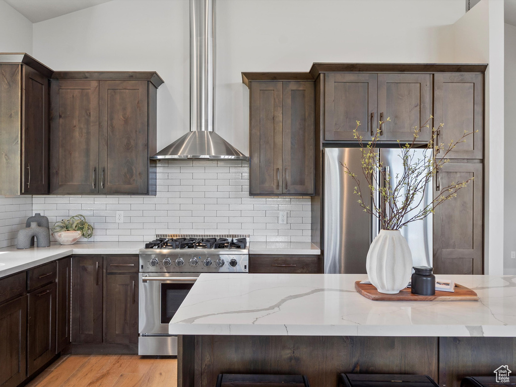 Kitchen with appliances with stainless steel finishes, wall chimney range hood, light stone countertops, tasteful backsplash, and dark brown cabinets