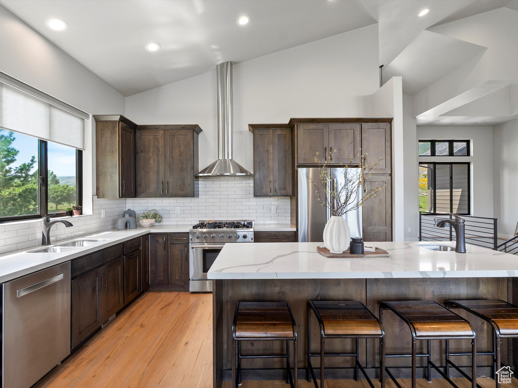 Kitchen with appliances with stainless steel finishes, lofted ceiling, a sink, wall chimney exhaust hood, and light stone countertops