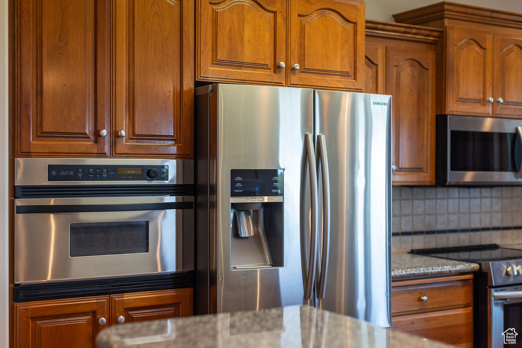 Kitchen featuring appliances with stainless steel finishes, light stone countertops, decorative backsplash, and brown cabinets