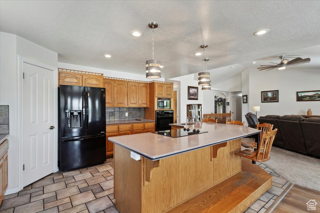 Kitchen featuring black appliances, open floor plan, light countertops, an island with sink, and a breakfast bar area