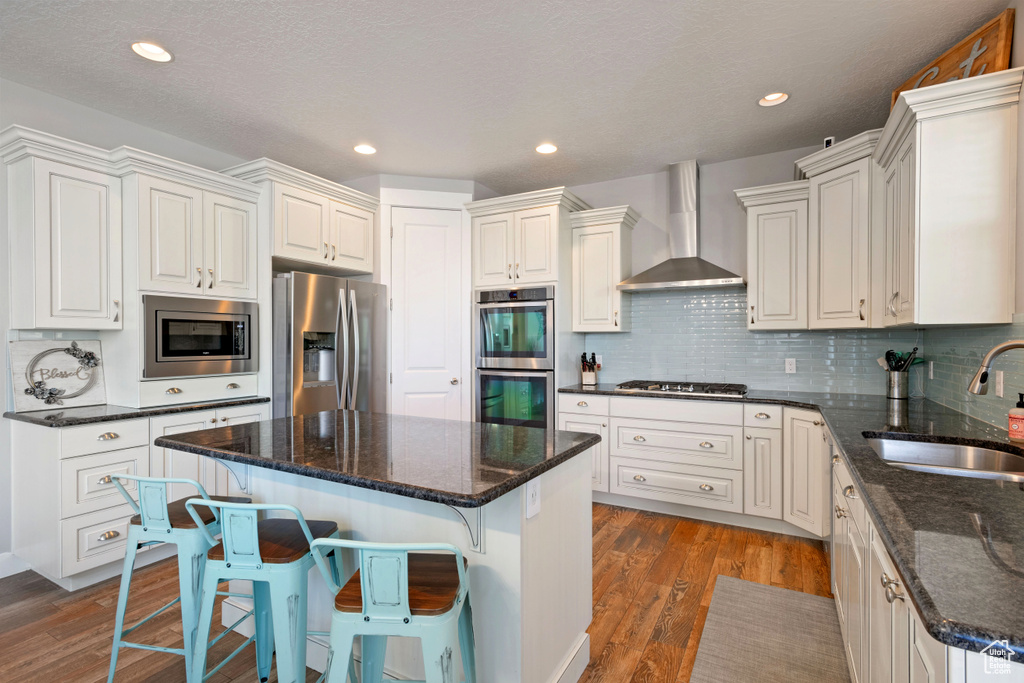 Kitchen featuring wall chimney range hood, a sink, stainless steel appliances, a kitchen island, and wood finished floors