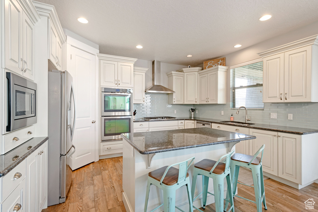 Kitchen featuring a sink, appliances with stainless steel finishes, wall chimney exhaust hood, a breakfast bar area, and light wood-style flooring