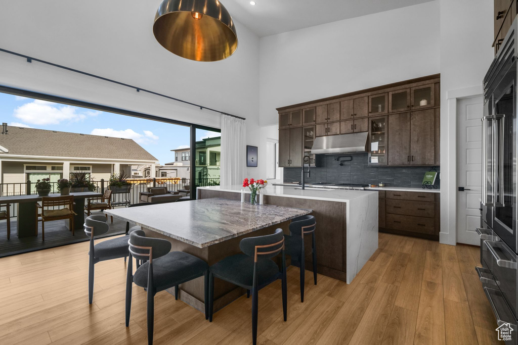 Kitchen featuring a towering ceiling, under cabinet range hood, dark brown cabinetry, light wood-style floors, and an island with sink