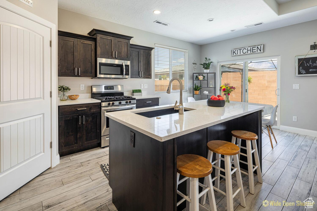 Kitchen with appliances with stainless steel finishes, a sink, light wood-style floors, a kitchen bar, and an island with sink