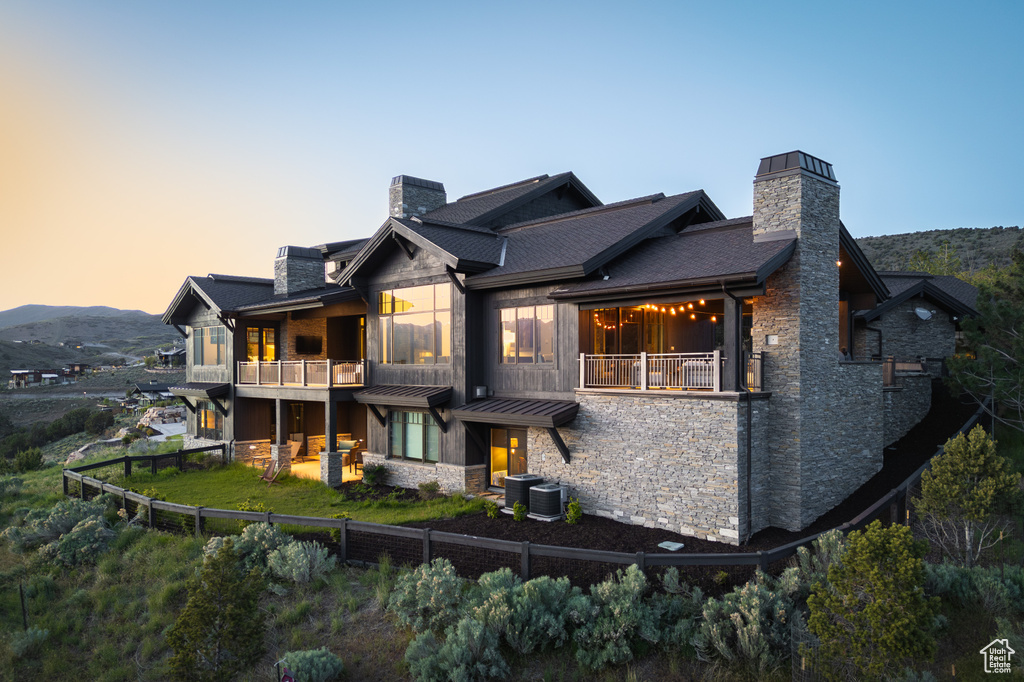 Back of house at dusk featuring a chimney, stone siding, a balcony, a fenced backyard, and central AC unit