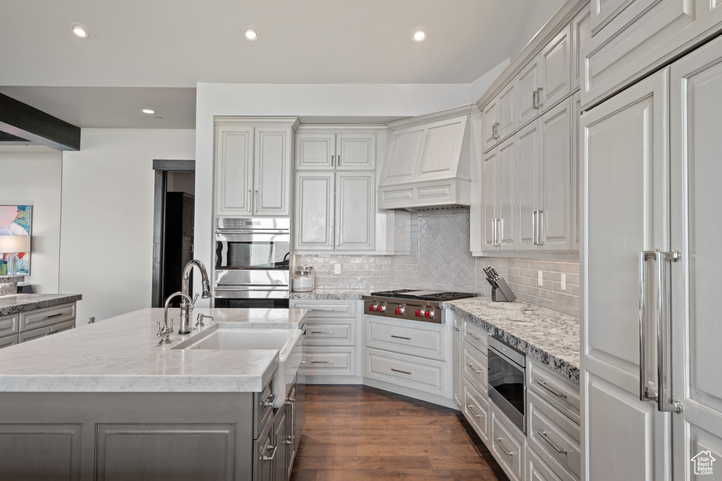 Kitchen with premium range hood, stainless steel appliances, decorative backsplash, a kitchen island with sink, and gray cabinetry