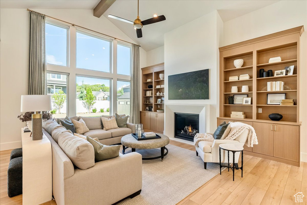 Living area featuring light wood finished floors, beamed ceiling, a warm lit fireplace, high vaulted ceiling, and ceiling fan