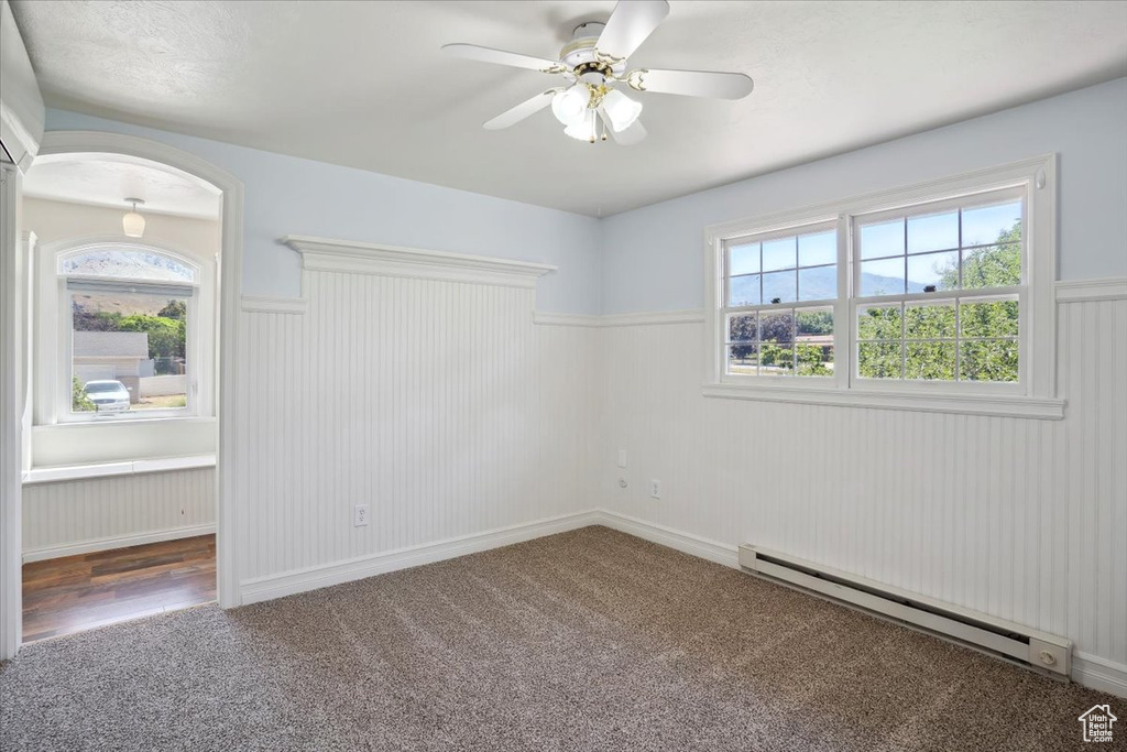 Carpeted spare room with a baseboard radiator, wainscoting, a ceiling fan, plenty of natural light, and arched walkways