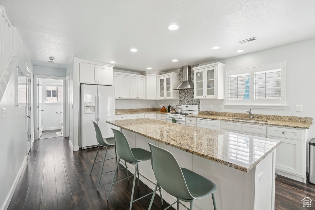 Kitchen featuring white appliances, wall chimney range hood, a kitchen breakfast bar, a sink, and white cabinetry