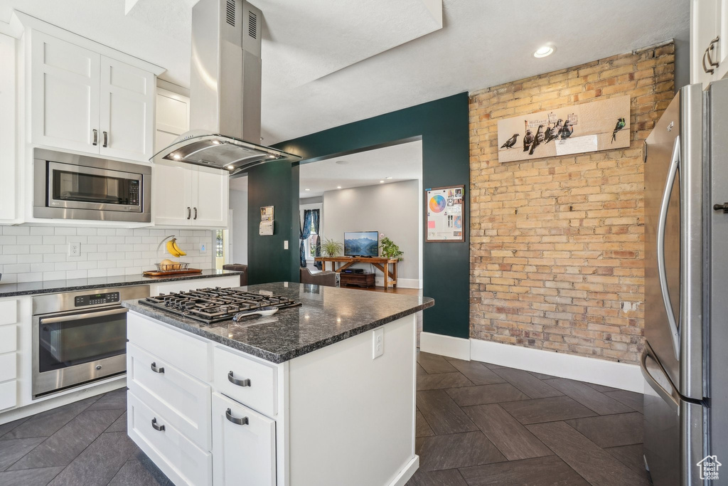 Kitchen with appliances with stainless steel finishes, island exhaust hood, baseboards, white cabinetry, and brick wall