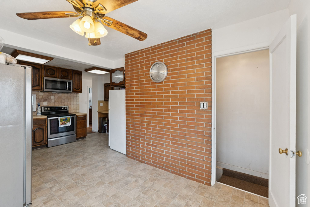 Kitchen with appliances with stainless steel finishes, a ceiling fan, dark brown cabinets, stone finish flooring, and brick wall