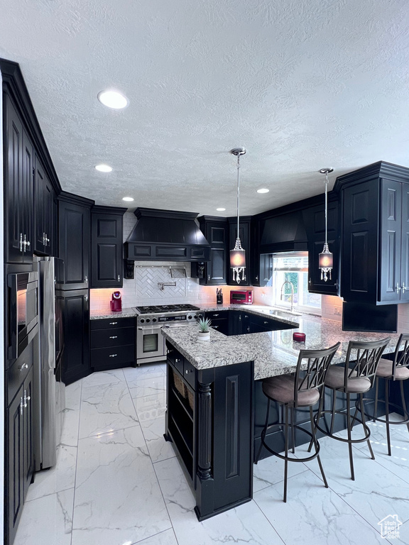 Kitchen with appliances with stainless steel finishes, custom range hood, light marble finish flooring, dark cabinetry, and light stone counters