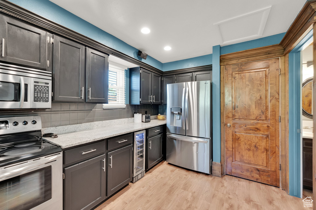 Kitchen with appliances with stainless steel finishes, wine cooler, decorative backsplash, light wood-style floors, and light stone countertops