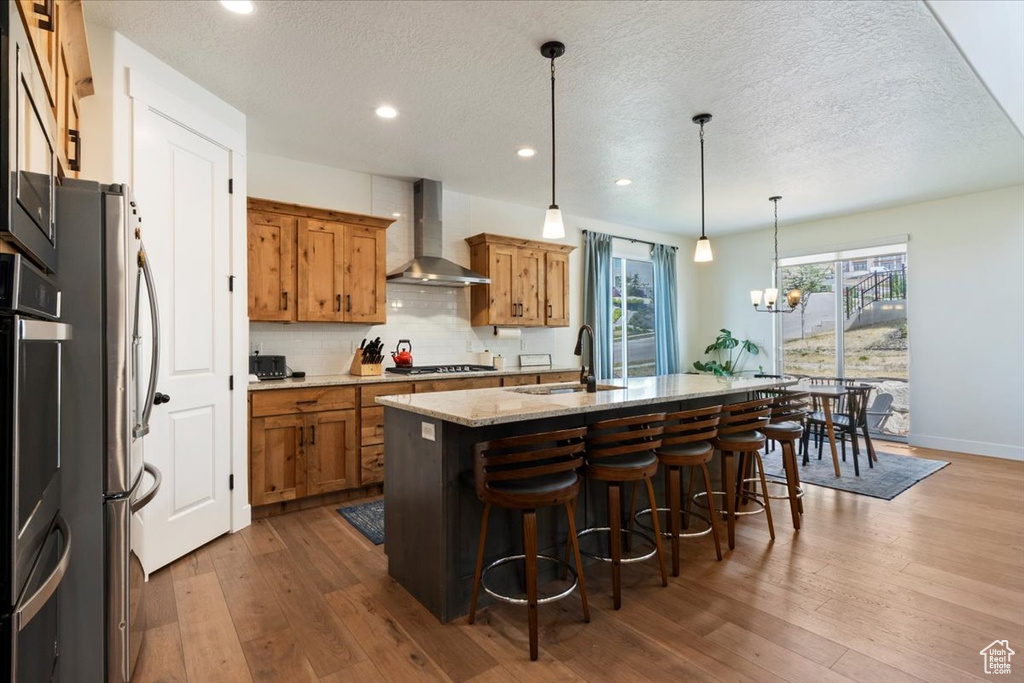 Kitchen featuring wall chimney exhaust hood, brown cabinetry, stainless steel appliances, a sink, and a kitchen island with sink