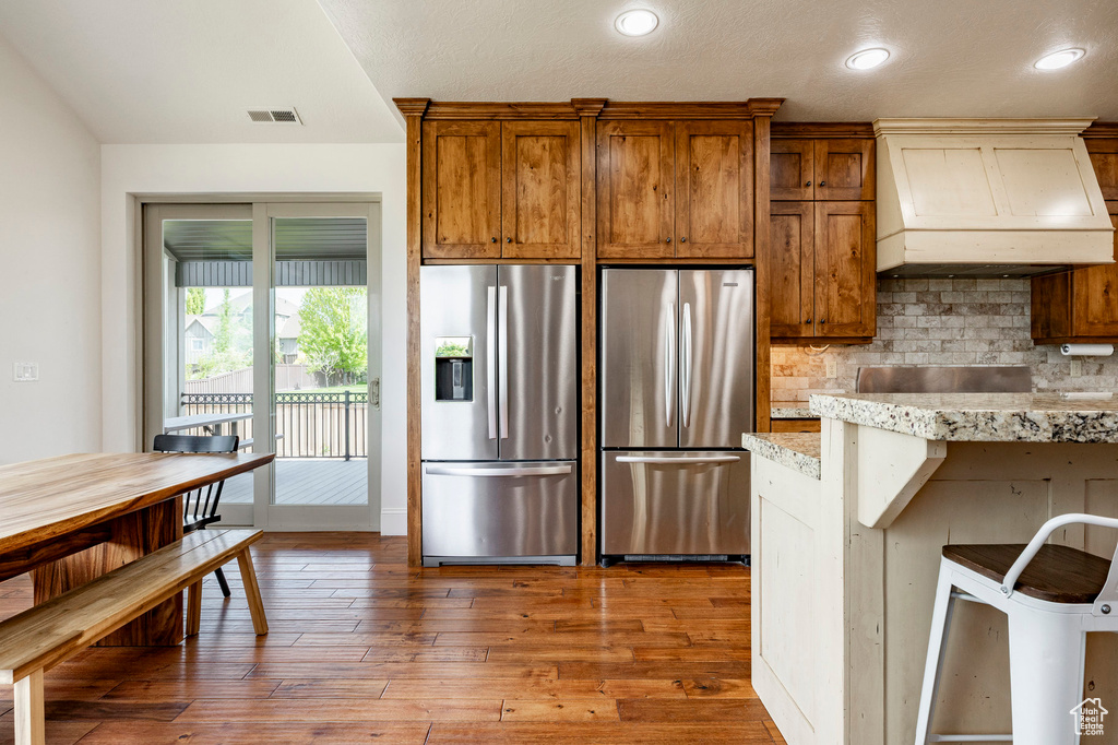 Kitchen with stainless steel refrigerator with ice dispenser, freestanding refrigerator, custom range hood, decorative backsplash, and hardwood / wood-style floors