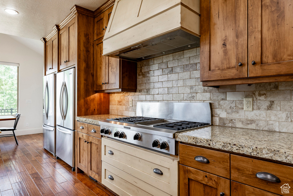 Kitchen with extractor fan, appliances with stainless steel finishes, brown cabinetry, decorative backsplash, and vaulted ceiling