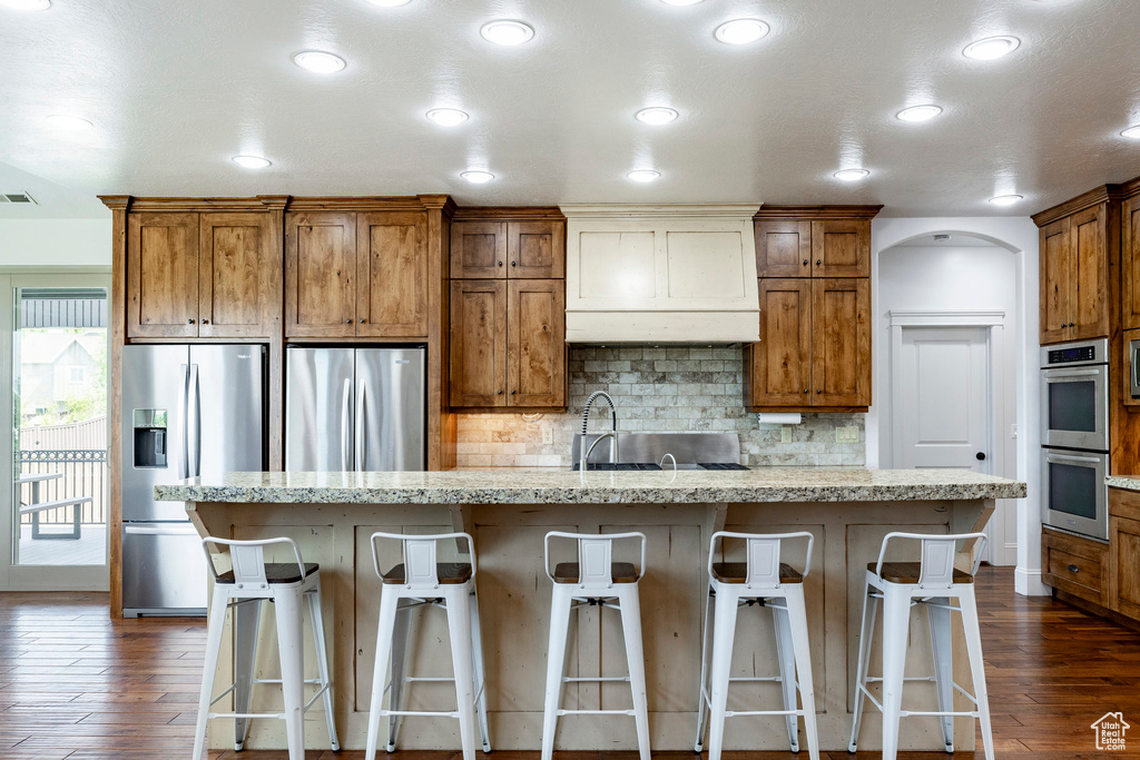Kitchen with stainless steel appliances, dark wood finished floors, brown cabinetry, backsplash, and an island with sink