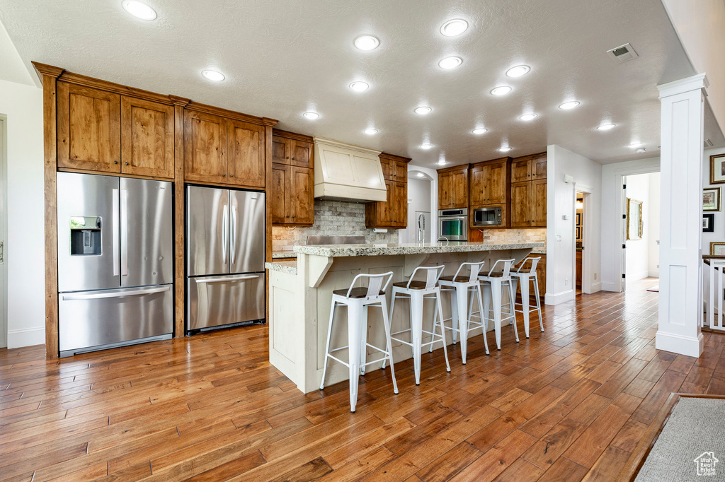 Kitchen with stainless steel appliances, custom exhaust hood, light wood-style floors, brown cabinets, and backsplash