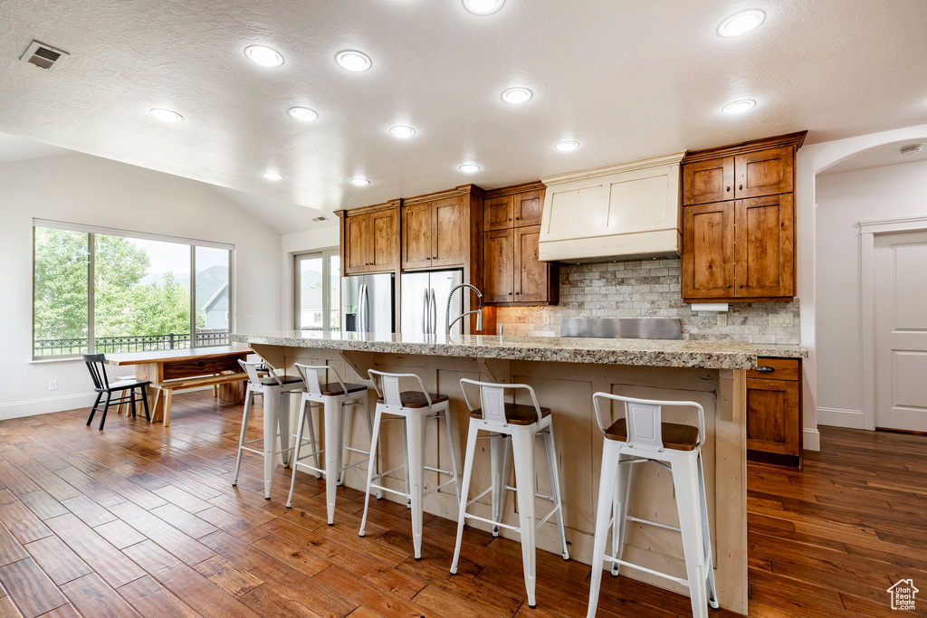 Kitchen featuring premium range hood, brown cabinets, dark wood-style floors, vaulted ceiling, and backsplash