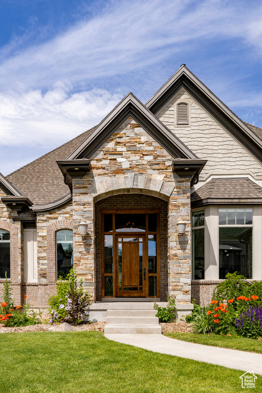 View of exterior entry with roof with shingles, a yard, brick siding, and stone siding