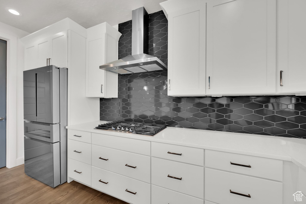 Kitchen featuring wall chimney range hood, stainless steel appliances, white cabinetry, light wood finished floors, and backsplash