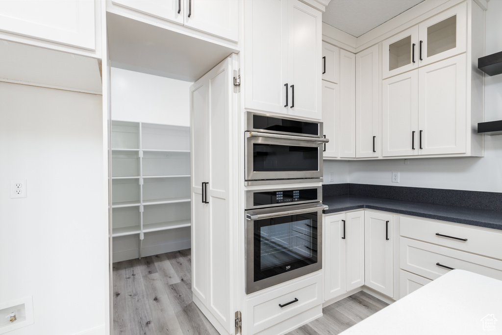 Kitchen with stainless steel double oven, light wood finished floors, white cabinetry, and dark countertops