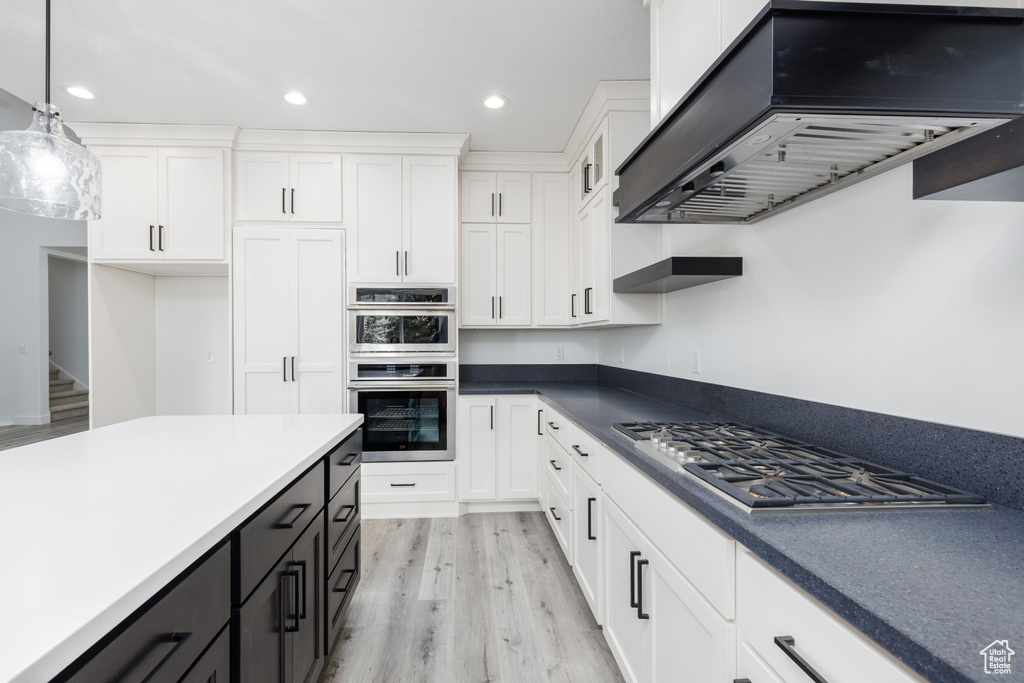 Kitchen featuring wall chimney range hood, stainless steel appliances, white cabinets, light wood-style floors, and hanging light fixtures
