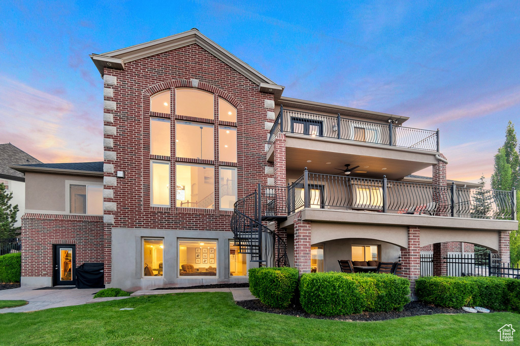 Back of property at dusk featuring a balcony, brick siding, a yard, stucco siding, and ceiling fan