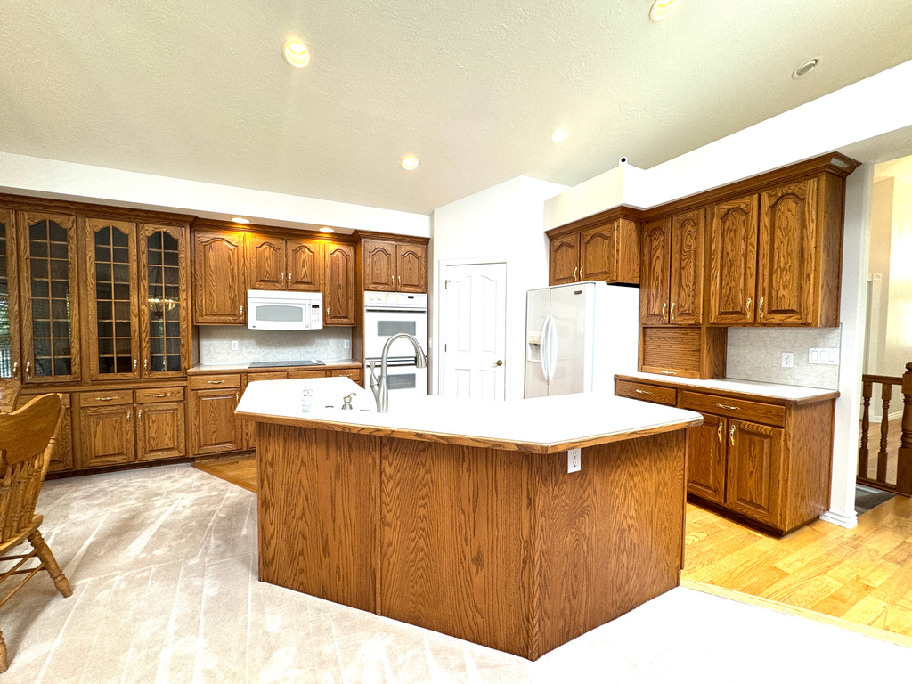 Kitchen featuring white appliances, light countertops, brown cabinetry, an island with sink, and recessed lighting