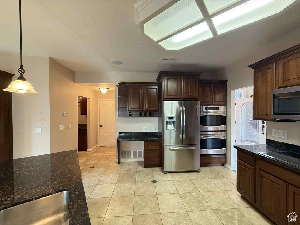 Kitchen with stainless steel appliances, dark brown cabinetry, dark stone counters, pendant lighting, and light tile patterned floors