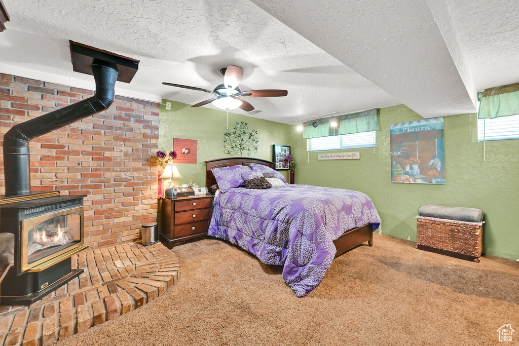 Carpeted bedroom with a wood stove, a textured ceiling, multiple windows, ceiling fan, and a textured wall