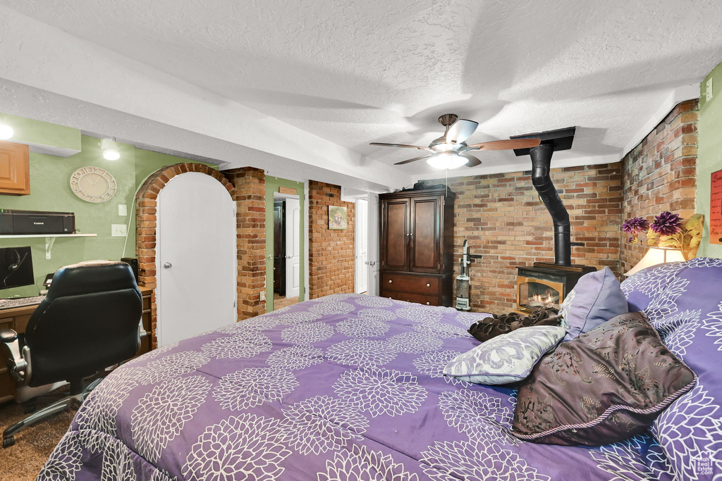 Carpeted bedroom featuring brick wall, a wood stove, a textured ceiling, ceiling fan, and an office area