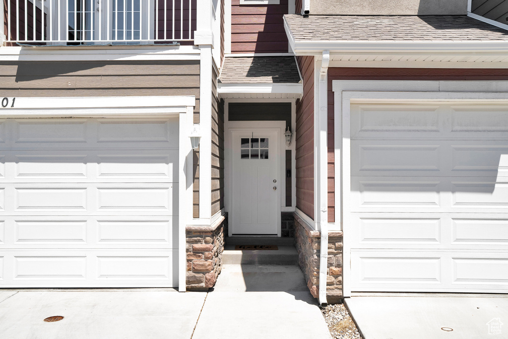 View of exterior entry with stone siding, roof with shingles, a balcony, concrete driveway, and a garage