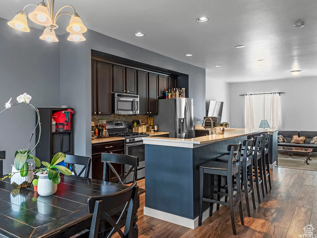 Kitchen featuring stainless steel appliances, dark wood-style flooring, dark brown cabinetry, a kitchen island with sink, and backsplash