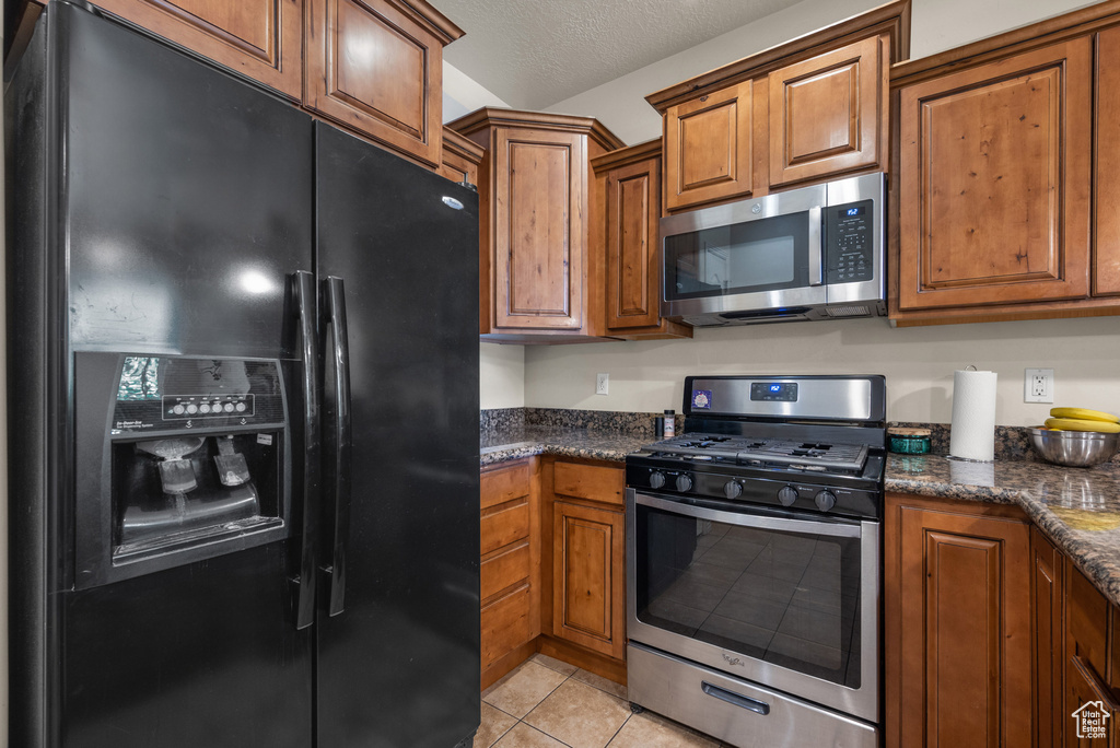 Kitchen with stainless steel appliances, brown cabinetry, light tile patterned floors, a textured ceiling, and dark stone countertops