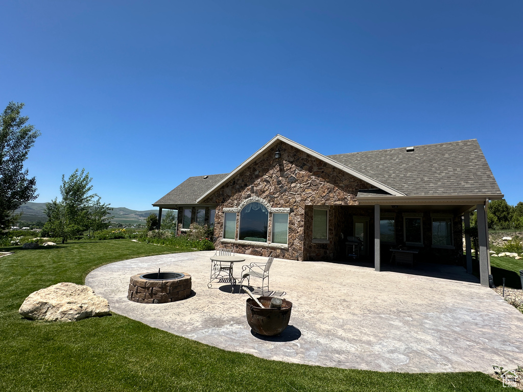 Rear view of house featuring a fire pit, a patio, a yard, stone siding, and roof with shingles