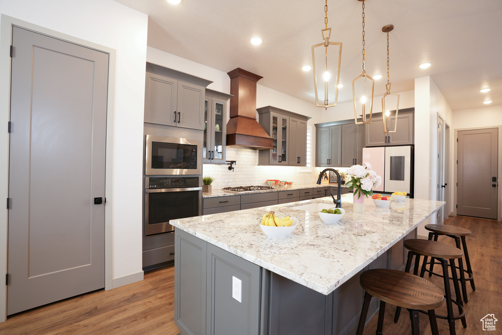 Kitchen featuring stainless steel appliances, custom exhaust hood, gray cabinetry, backsplash, and a kitchen bar